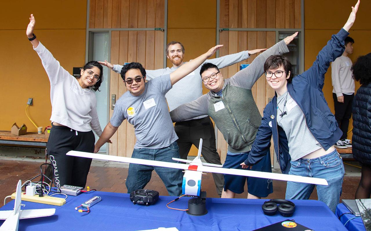 Five students with their arms out like they are flying. They're standing in front of a model plane.